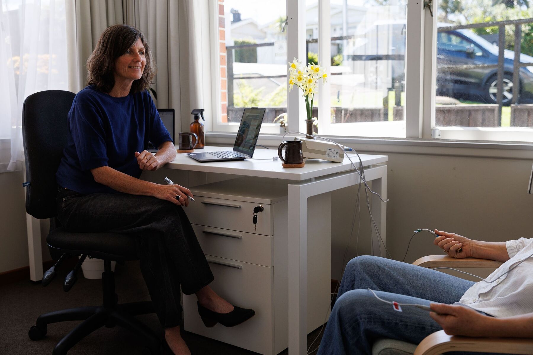 Caroline smiling at her desk by a sunny window during a bioresonance session, with the client seated in the foreground holding the hand electrodes
