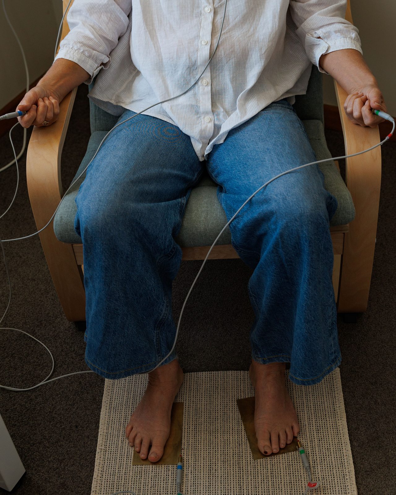 A client seated in the studio armchair holding the hand electrodes, with her bare feet resting on brass foot plates