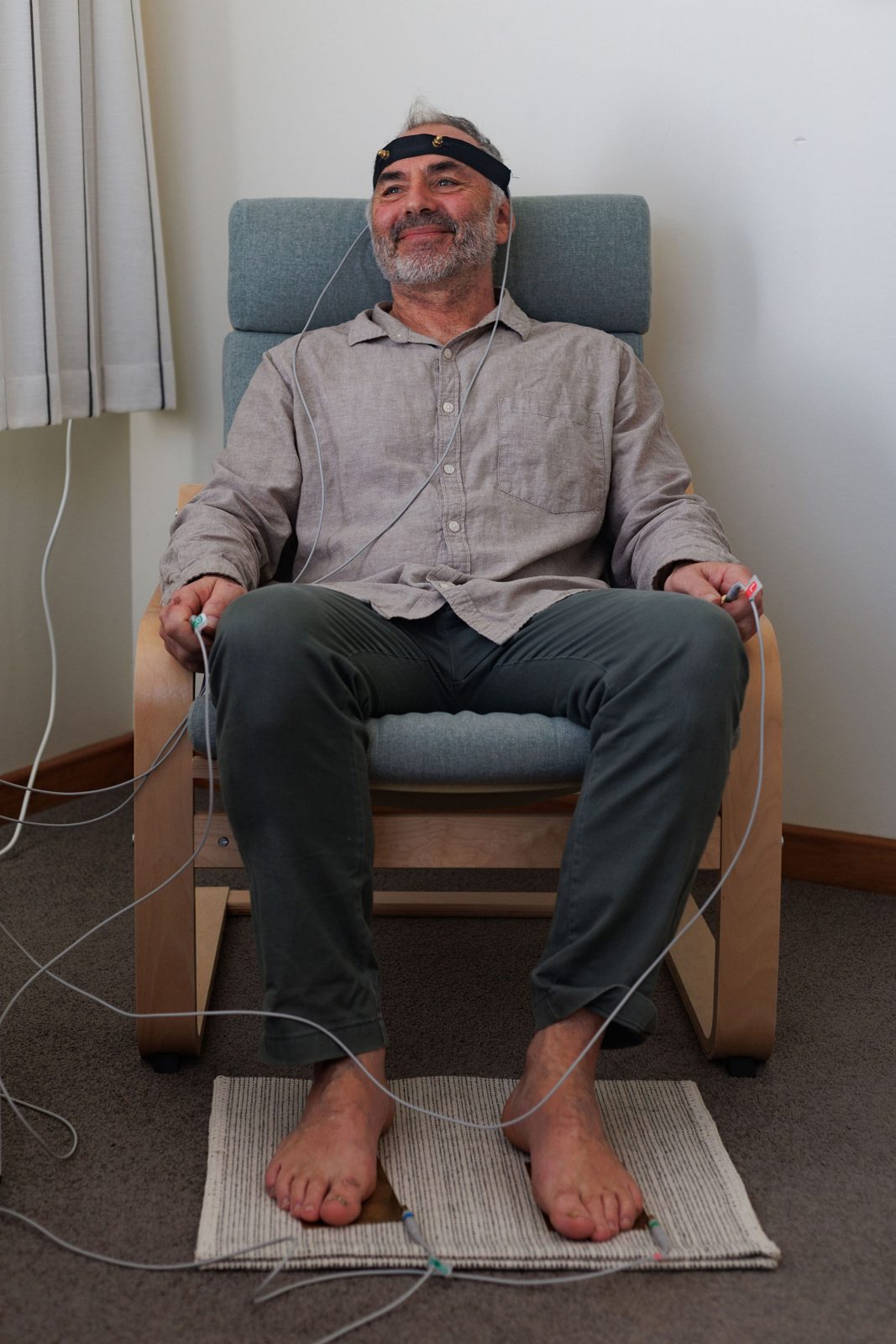 A client smiling in the studio armchair during a bioresonance session, wearing the headband and holding the hand electrodes